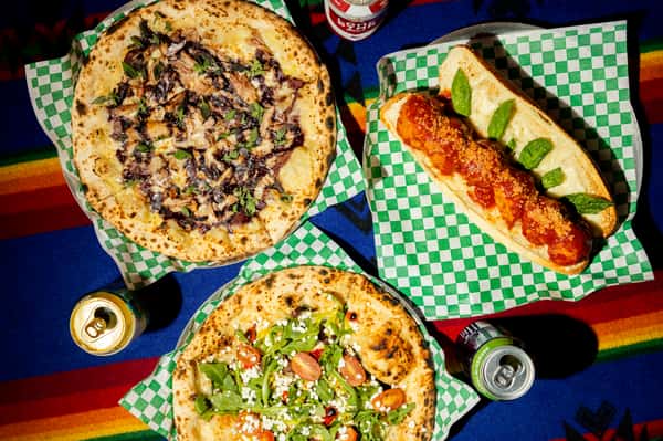 Wood-fired pizzas with gourmet toppings and a meatball sub with marinara, served with canned drinks on a colorful tablecloth.