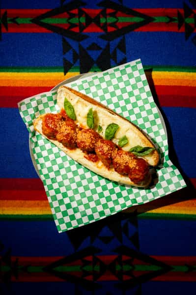 Meatball sub with marinara sauce and grated cheese on toasted bread, garnished with fresh basil on checkered paper.