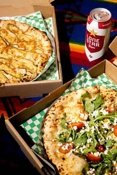 Wood-fired pizzas in takeout boxes—one with arugula and cherry tomatoes, one with sliced vegetables—served with canned beer on colorful cloth.