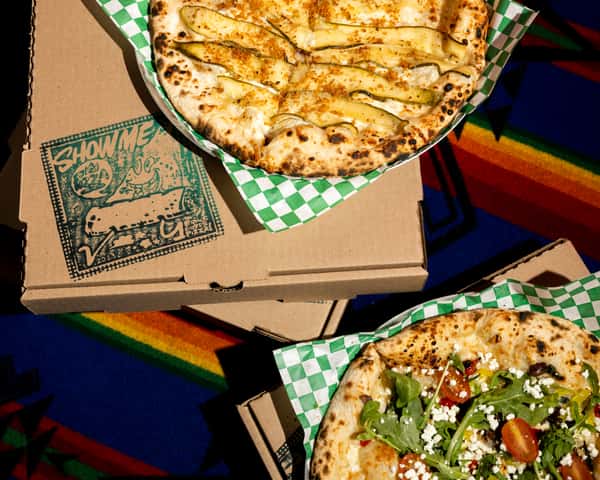 Wood-fired pizzas in takeout boxes—one with sliced zucchini and cheese, one with arugula and tomatoes—on a colorful tablecloth.