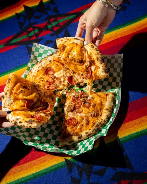Wood-fired pizza topped with pepperoni, sausage, and melted cheese on a blistered crust over colorful textile backdrop