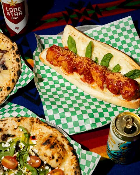 Meatball sub with marinara, melted cheese, and basil on toasted roll, served with artisan pizzas and canned drinks.
