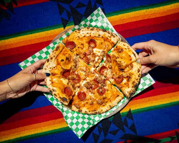 Wood-fired pizza topped with pepperoni, sausage, and melted cheese on a blistered crust over colorful textile backdrop