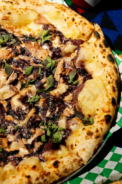 Close-up of wood-fired pizza topped with melted cheese, shredded meat, herbs, and rich dark sauce on blistered crust.
