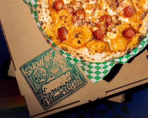 Wood-fired pizza topped with pepperoni, sausage, and melted cheese on a blistered crust over colorful textile backdrop