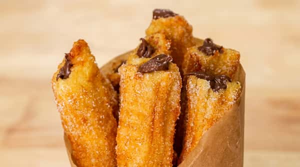 Golden-brown churros dusted with sugar, topped with dollops of melted chocolate, served in a brown paper cone.