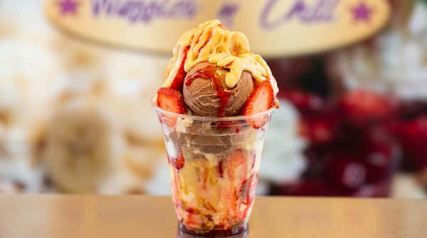Ice cream sundae with chocolate and strawberry scoops, sliced strawberries, and yellow/red drizzles, in a clear cup.