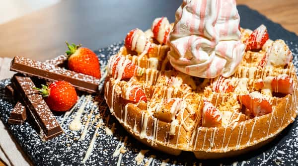 Waffle topped with whipped cream, strawberries, white chocolate drizzle, and chocolate bars on a dark surface.