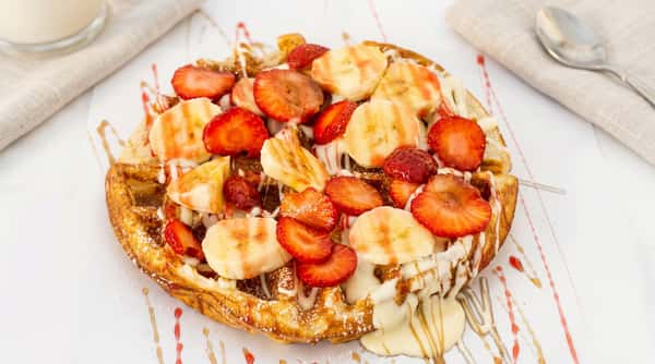 Round waffle topped with sliced strawberries and bananas, drizzled with white chocolate and caramel.