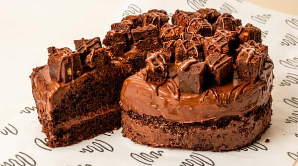 Chocolate fudge cake with a slice removed, topped with brownie pieces and a drizzle of chocolate sauce.