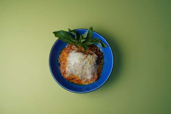 Baked lasagna with melted cheese and basil leaves served in a blue bowl on a green background