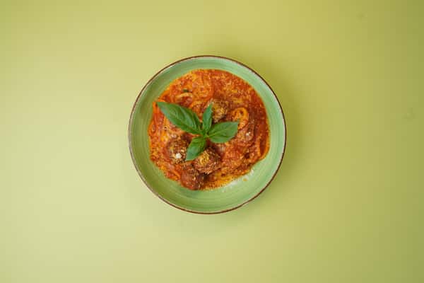 Overhead view of spaghetti with meatballs in tomato sauce, served with basil in a green bowl on a green background