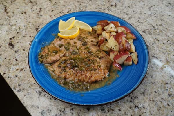 Chicken piccata garnished with lemon slices and served with roasted red potatoes on a blue plate over a granite countertop