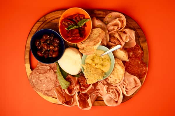 Italian antipasto charcuterie board with salami, prosciutto, mortadella, mozzarella, olives, and sun-dried tomatoes on orange background.