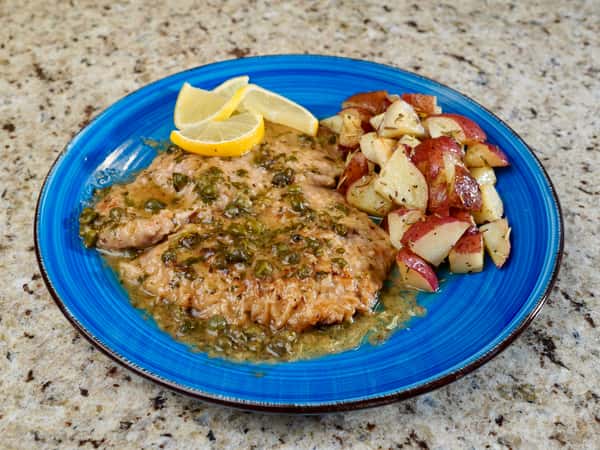 Lemon chicken piccata with capers served alongside roasted red potatoes on a blue plate
