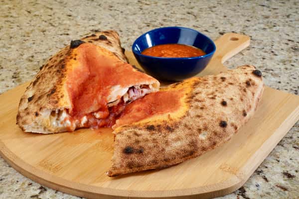 Wood-fired calzone filled with cheese and ham, topped with tomato sauce and served with a side of marinara in a blue bowl