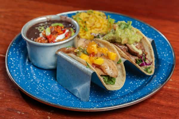 Two tacos filled with grilled meat, mango salsa, guacamole, and slaw, served with black beans and yellow rice on a blue plate.
