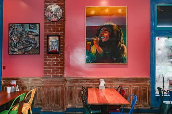 Vibrant restaurant interior with multicolored chairs, wooden tables, a reggae singer painting, and large windows facing the street.