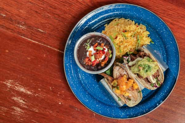 Two tacos with mango salsa and guacamole, served with black beans, yellow rice, and garnished with scallions on a blue speckled plate.