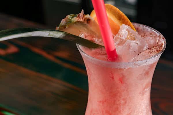 Pink frozen cocktail topped with ice, garnished with a pineapple slice and orange wedge, served with a red straw.