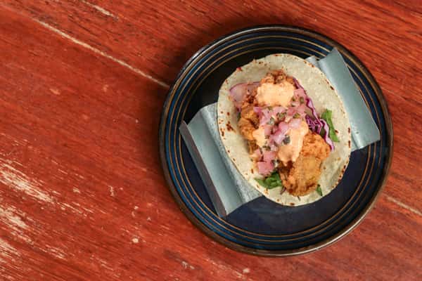 Fried chicken taco topped with pink onions, cabbage, and creamy sauce on a corn tortilla, served on a blue-rimmed plate.