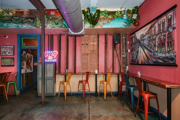 Colorful dining area with rainbow metal chairs, wooden tables, tropical wall art, and a TV showing a basketball game.