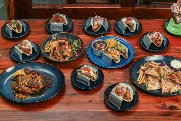 Assorted Mexican-inspired dishes on a wooden table, featuring tacos, quesadillas, grilled chicken, rice, beans, and a dessert parfait.
