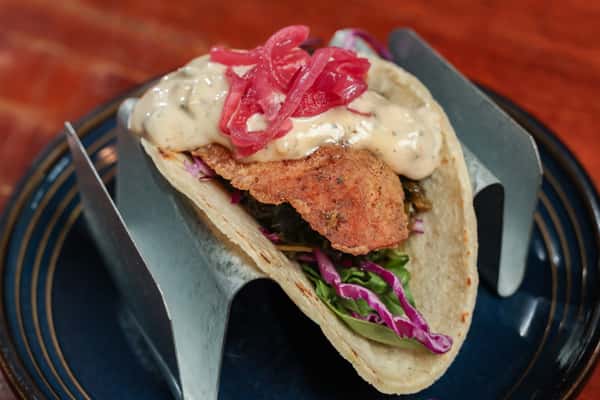 Crispy fish taco topped with creamy sauce, pickled red onions, shredded cabbage, and greens in a soft tortilla shell.