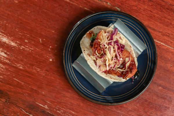 Crispy fried fish taco topped with purple cabbage slaw and a drizzle of tangy sauce, served on a soft corn tortilla.
