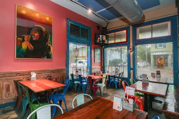 Vibrant restaurant interior with multicolored chairs, wooden tables, a reggae singer painting, and large windows facing the street.