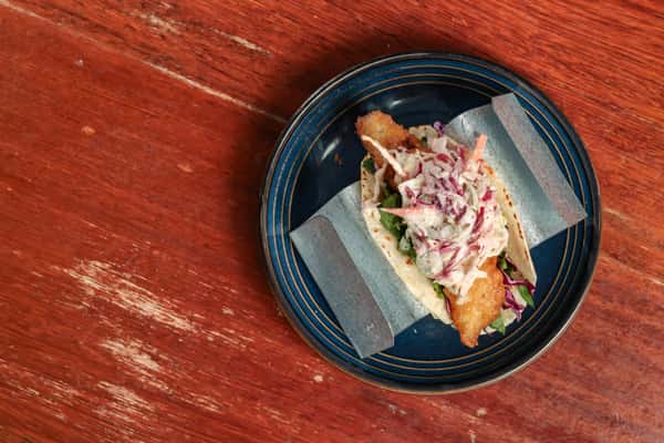 Crispy fried fish taco topped with creamy coleslaw and served in a flour tortilla on a blue-rimmed plate.