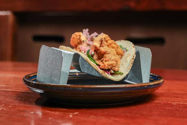 Fried chicken taco topped with pink onions, cabbage, and creamy sauce on a corn tortilla, served on a blue-rimmed plate.