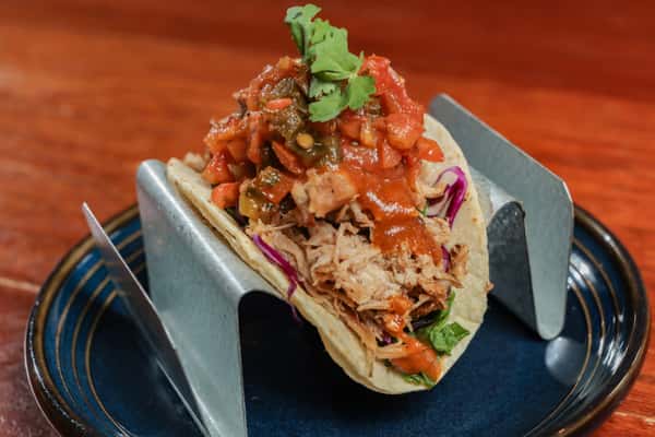 Pulled pork taco topped with fresh salsa and cilantro, served in a metal taco holder on a blue-rimmed plate.