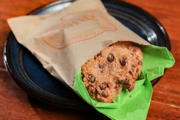 Chocolate chip cookie partially wrapped in a brown paper bag with green tissue paper, served on a blue-rimmed plate.