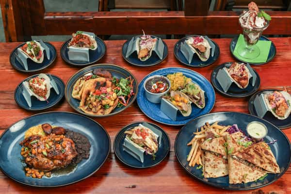 Assorted Carribean-inspired dishes on a wooden table, featuring tacos, quesadillas, grilled chicken, rice, beans, and a dessert parfait.