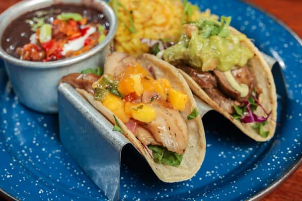 Two tacos served with mango salsa and guacamole, accompanied by black beans with toppings and yellow rice on a blue speckled plate.