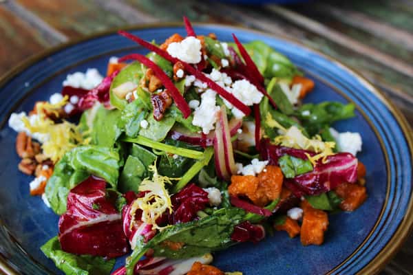 Fresh mixed greens salad with roasted sweet potatoes, goat cheese, beets, red onions, pecans, and lemon zest on a blue plate.