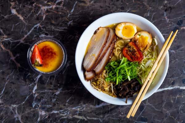 A Japanese ramen bowl with chashu pork, soft-boiled egg, and vegetables, served with chopsticks
