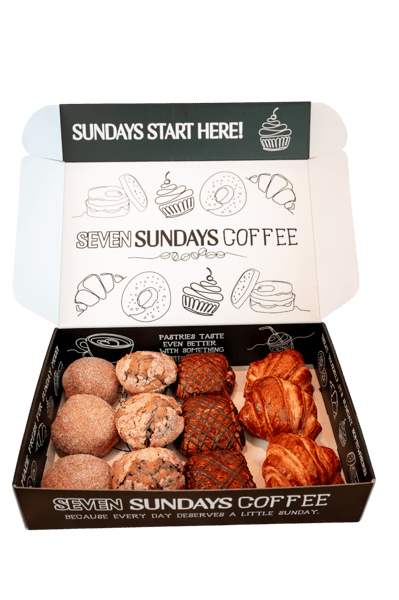 Open pastry box labeled "Seven Sundays Coffee," filled with assorted baked goods including croissants, Danish pastries with fruit filling, cookies, and other sweet pastries.