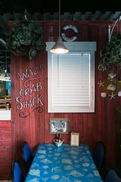 Rustic crab shack restaurant corner with red wooden walls, nautical décor, hanging plants, and a blue fish-patterned tablecloth.