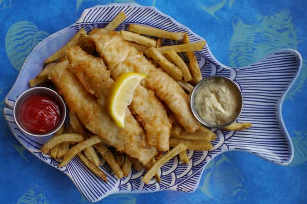 "Golden fried fish fillets with crispy French fries, served in a blue-and-white fish-shaped plate with tartar sauce, ketchup, and a fresh lemon wedge, set against a vibrant ocean-themed tablecloth.