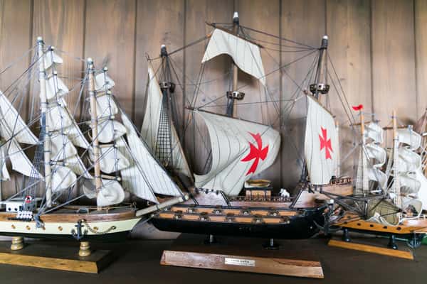 "Detailed model ships with sails and rigging, including a replica of a historic sailing vessel with red cross emblems, displayed against wooden paneling.