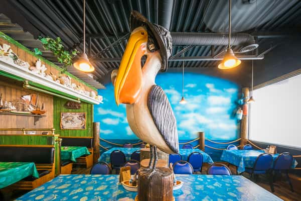 "Coastal-themed restaurant interior with a large wooden pelican statue centerpiece, blue ocean mural, and nautical-inspired seating.
