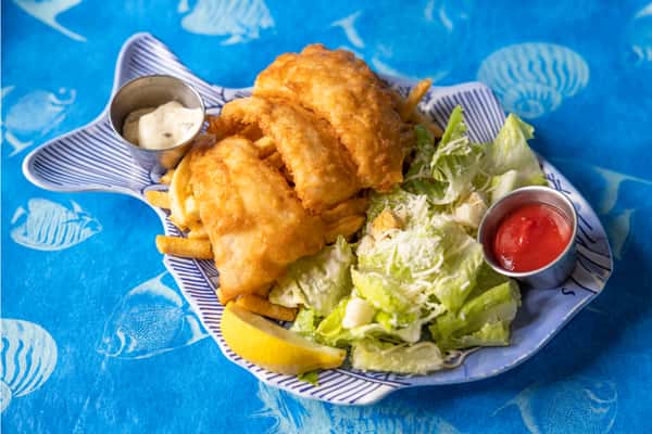 Beer-battered fish fillets with crispy fries, Caesar salad, lemon wedge, ketchup, and tartar sauce, plated on a fish-shaped dish with an ocean-themed background.