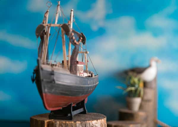 "Close-up of a detailed fishing boat model with red hull and intricate rigging, displayed against a blue ocean mural background with a seagull figurine.