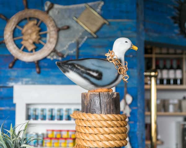 Decorative seagull statue perched on a wooden post wrapped in thick rope, set against a rustic blue wall with ship wheel and fishing net décor.