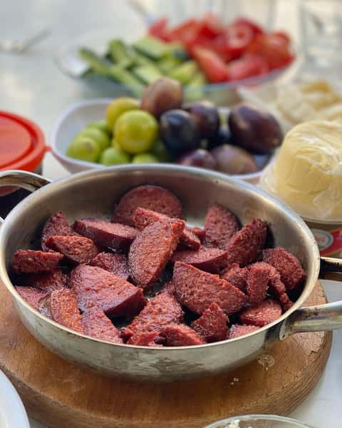 Traditional Turkish sucuk sausage sautéed in a hot skillet, served with fresh olives, tomatoes, cucumbers, and cheese for a classic Turkish breakfast at Ebby's Turkish Kitchen.