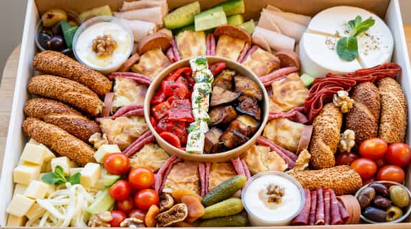 Charcuterie board with meats, cheeses, breads, a center bowl of roasted peppers and feta, olives, cherry tomatoes, and dips.