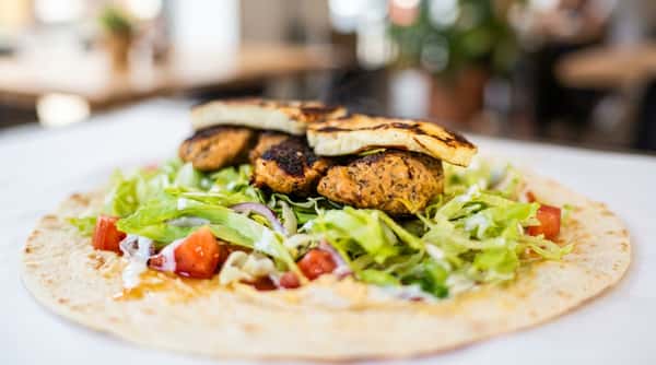 Steaming chicken meatballs and halloumi cheese on lettuce, red onion, tomatoes, and white sauce atop a tortilla.