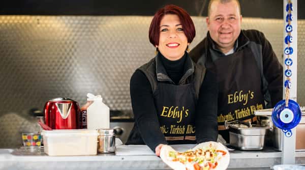 Woman in an "Ebby's Authentic Turkish Kitchen" apron holding a wrap, with a man smiling behind her.